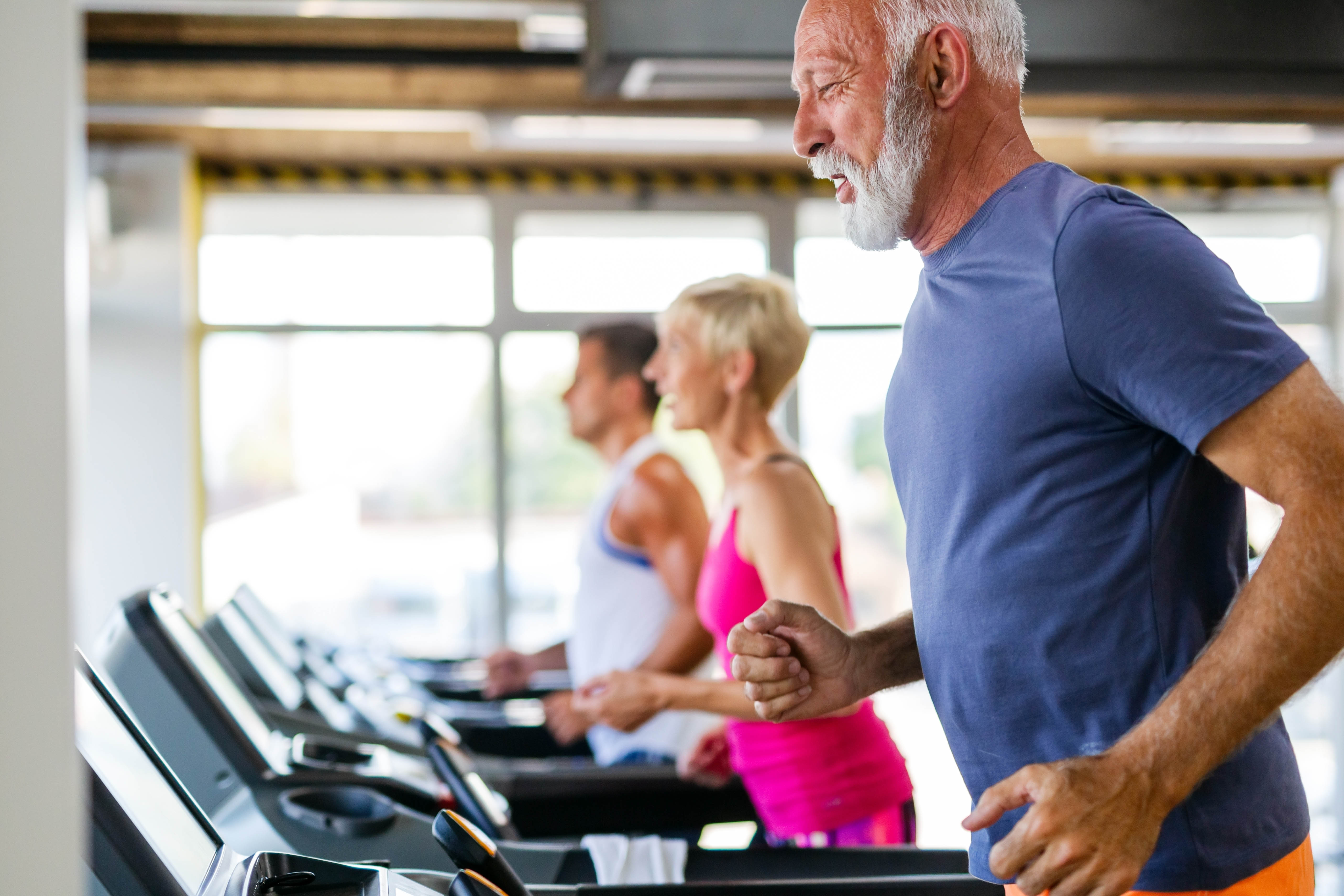 Older man running on a treadmill in a gym, with others in the background